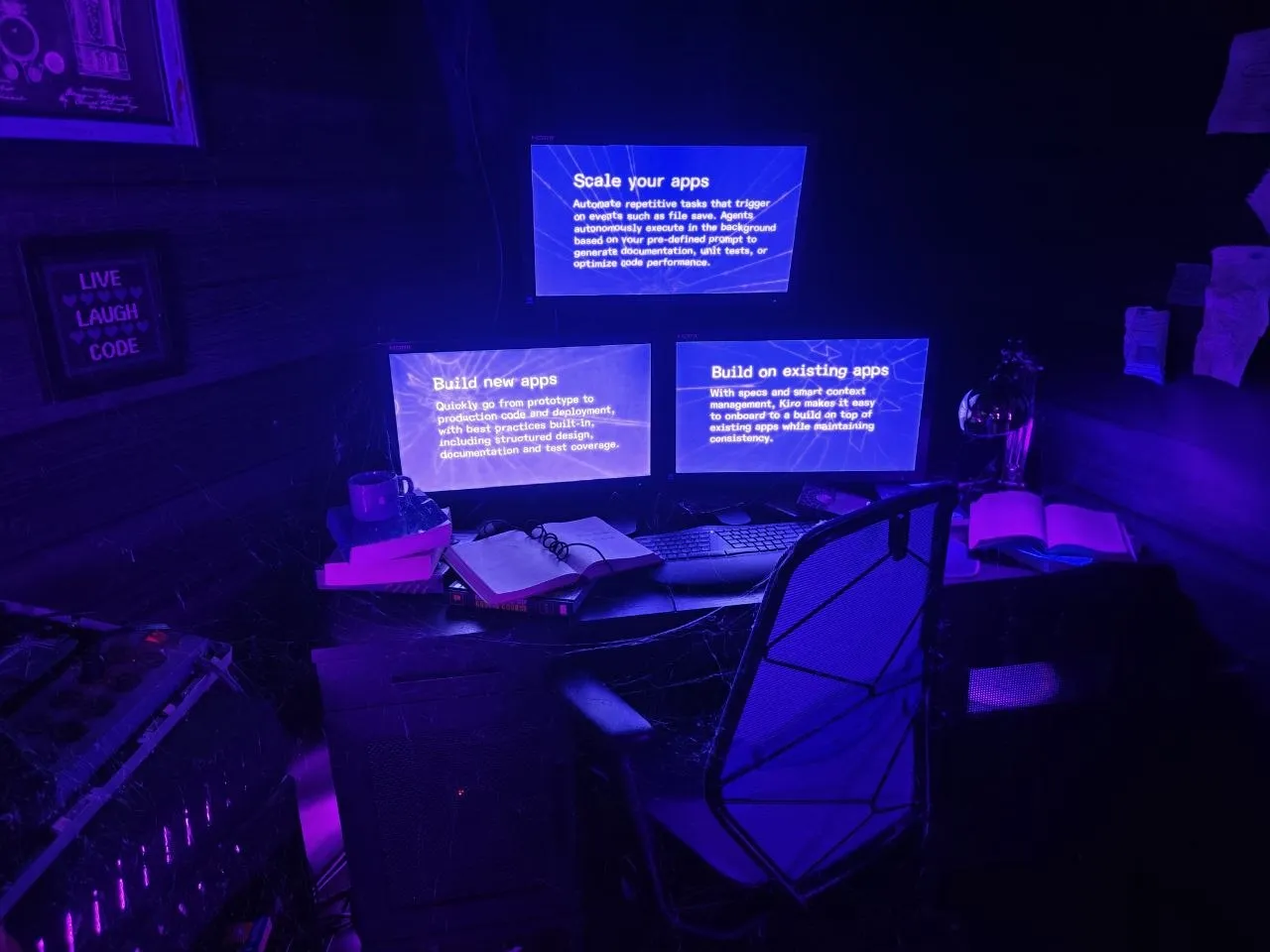 A photo taken from inside the House of Kiro, depicting a vacant, dilapidated desk with three monitors. Displayed upon them are Kiro's capabilities; building new apps, building upon existing apps, and scaling apps.
